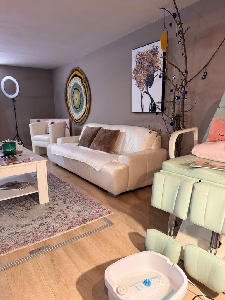 Ein geschmackvoll eingerichtetes Kosmetikstudio mit einem weißen Ledersofa und Sesseln, einem stilvollen Couchtisch, einem farbenfrohen abstrakten Gemälde an der Wand und einem dekorativen Zweig mit blauen Ornamenten. Im Vordergrund stehen ein Massagesessel und ein Fußbad.