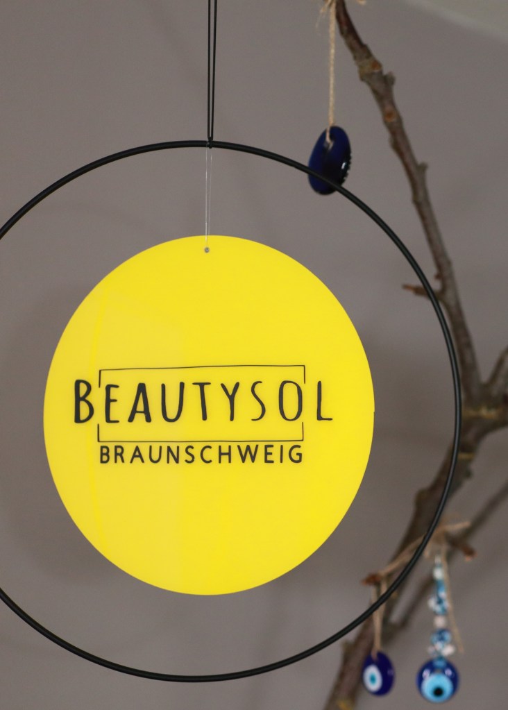 Ein rundes Schild mit dem Text „BEAUTYSOL BRAUNSCHWEIG“ in Schwarz auf leuchtend gelbem Hintergrund, aufgehängt in einem schwarzen runden Rahmen.