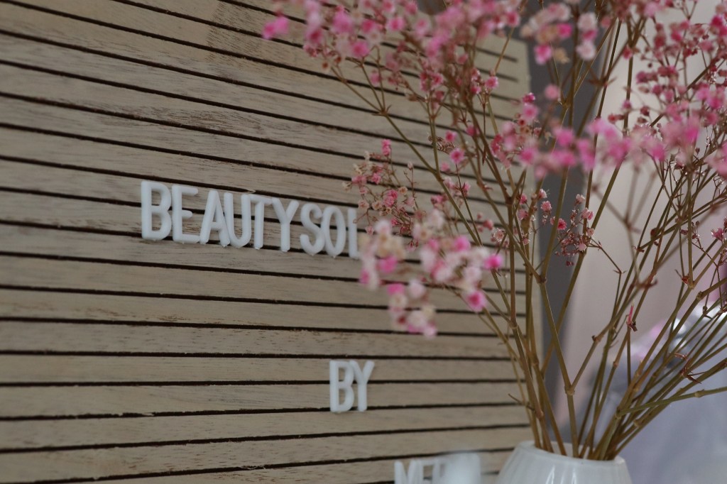 Eine Nahaufnahme einer dekorativen Wand mit den Worten „BEAUTYSOL BY MERAL“ in weißen Buchstaben, begleitet von einer Vase voller rosa Blumen vor einem Holzhintergrund.