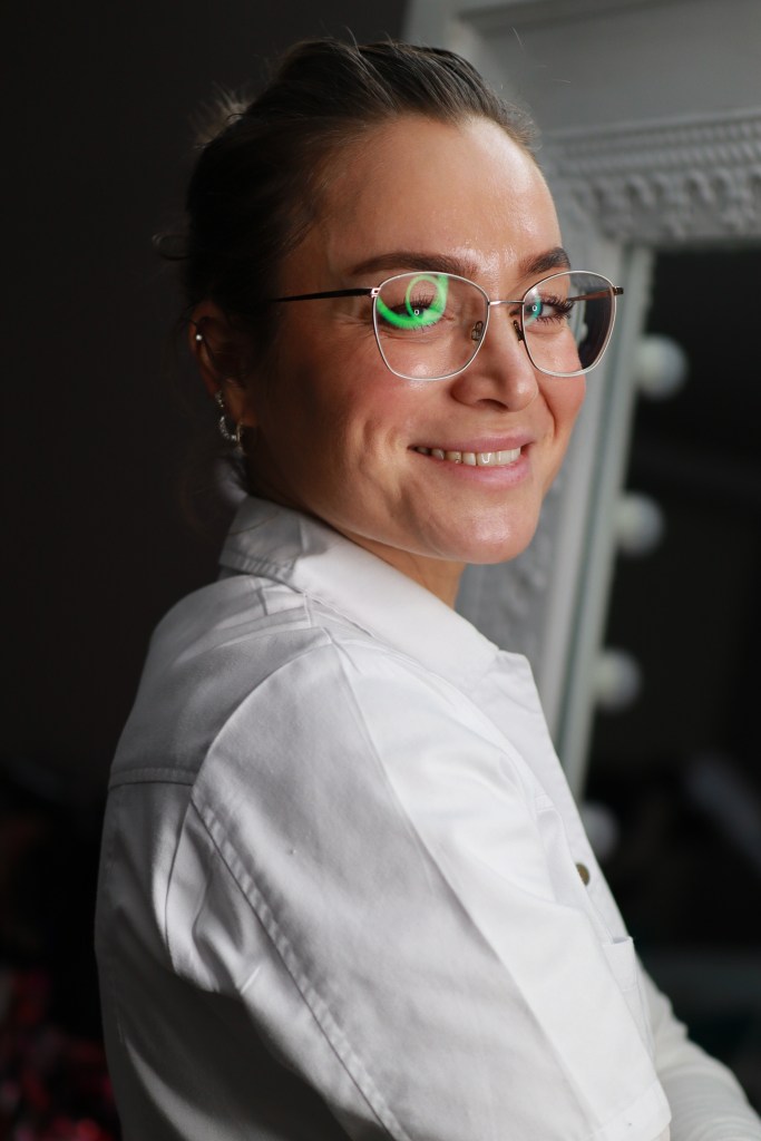 Frau mit Brille und weißem Kittel, lächelnd und vor einem Spiegel posierend.