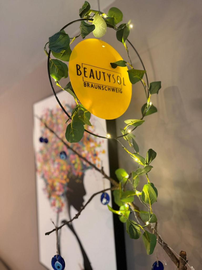 Ein leuchtend gelbes Schild mit der Aufschrift „BEAUTYSOL BRAUNSCHWEIG“, umgeben von Grün und Lichterketten, montiert auf einem Ast vor einem kunstvollen Gemälde.